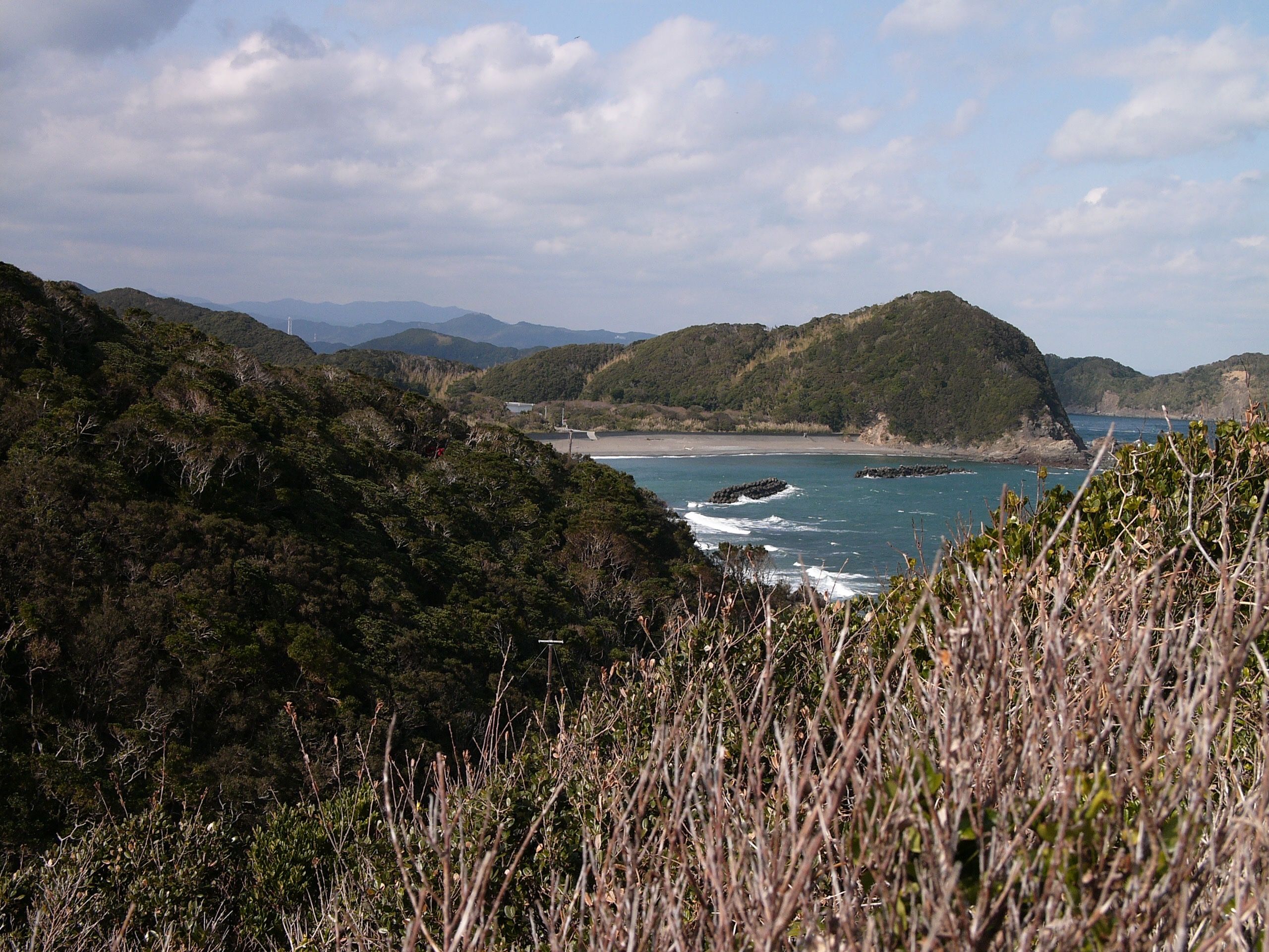 Panorama of a jagged seashore with two small bays and lines of forested hills.