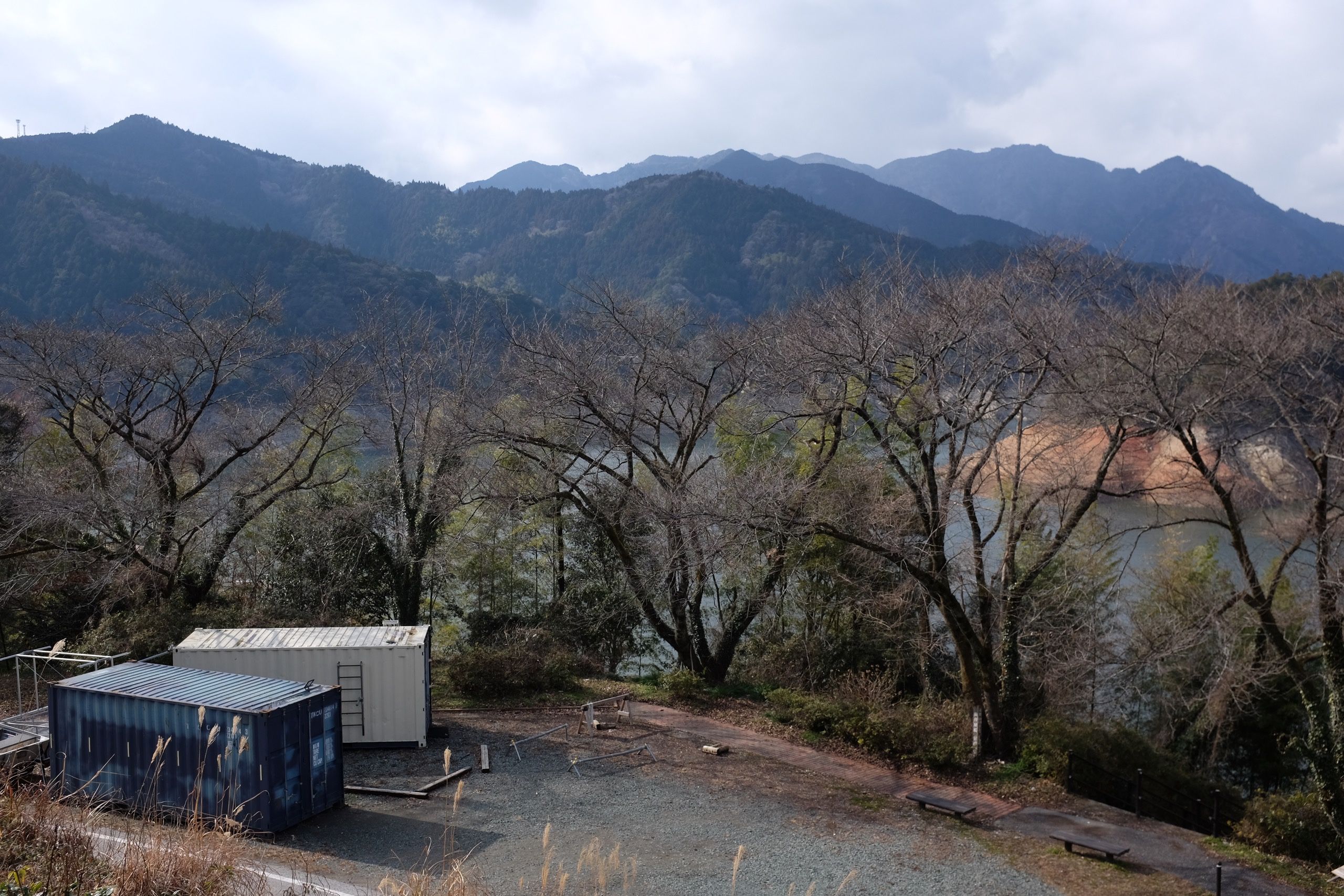 Looking down on two shipping containers in a parking lot in front of a row of large cherry trees. Behind is the artificial lake and the mountains.