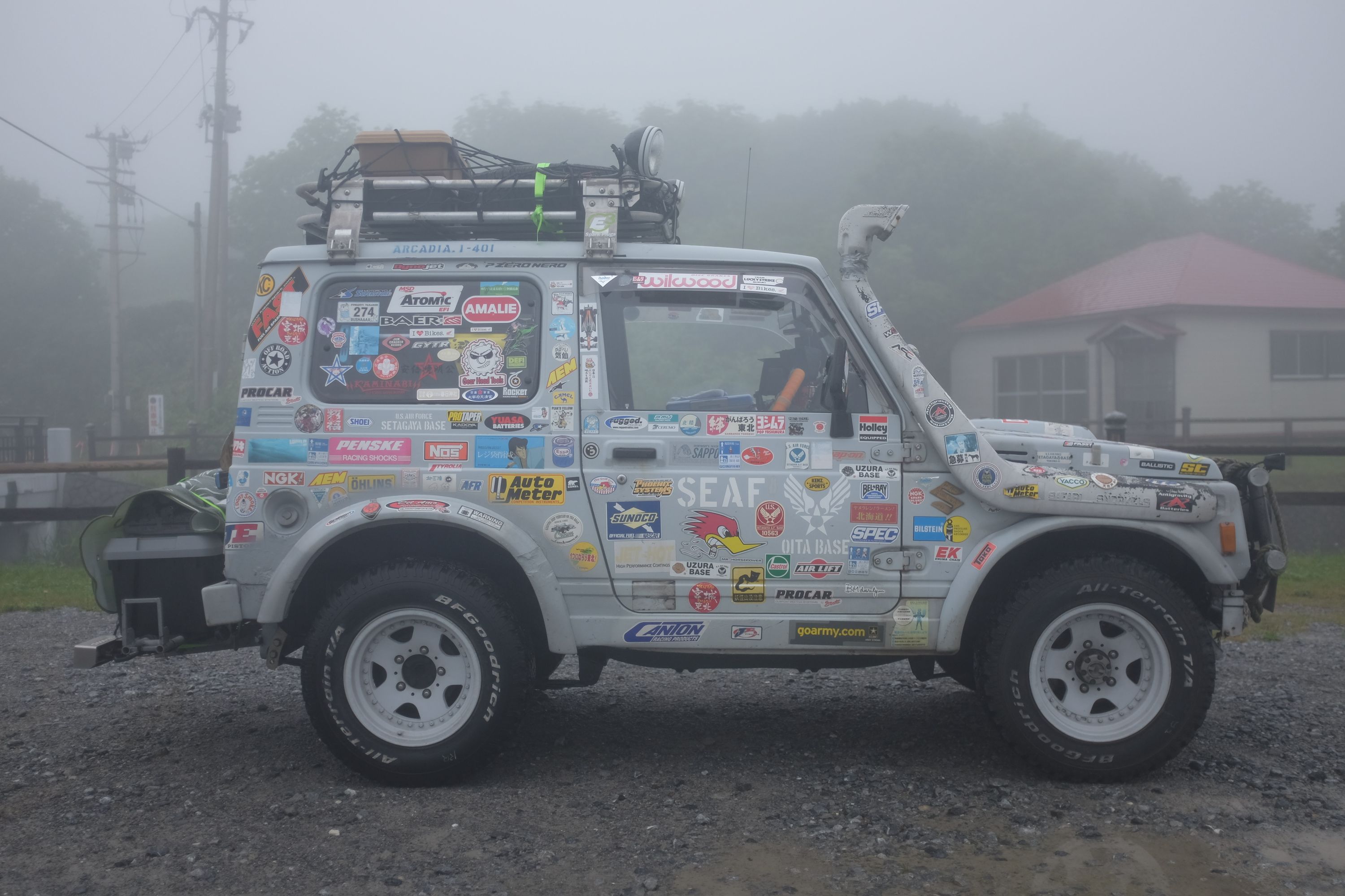 A small grey jeep festooned with stickers parked in the fog.