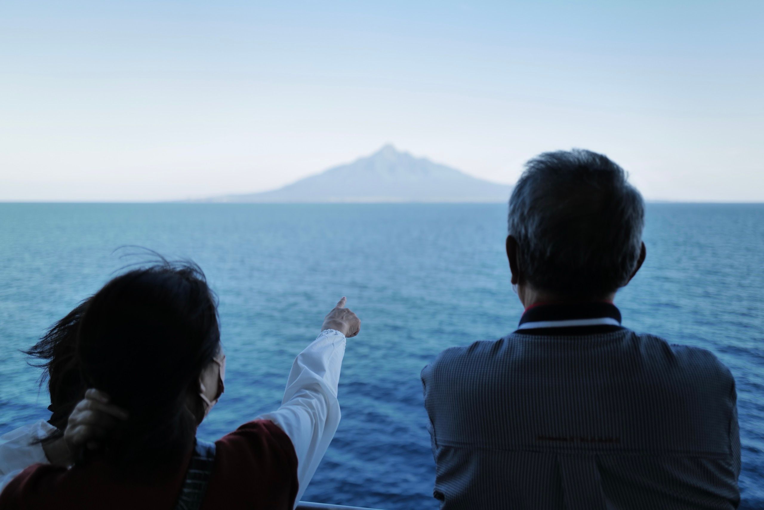 A woman, standing next to an older man, points at a volcano, Mount Rishiri, across the sea