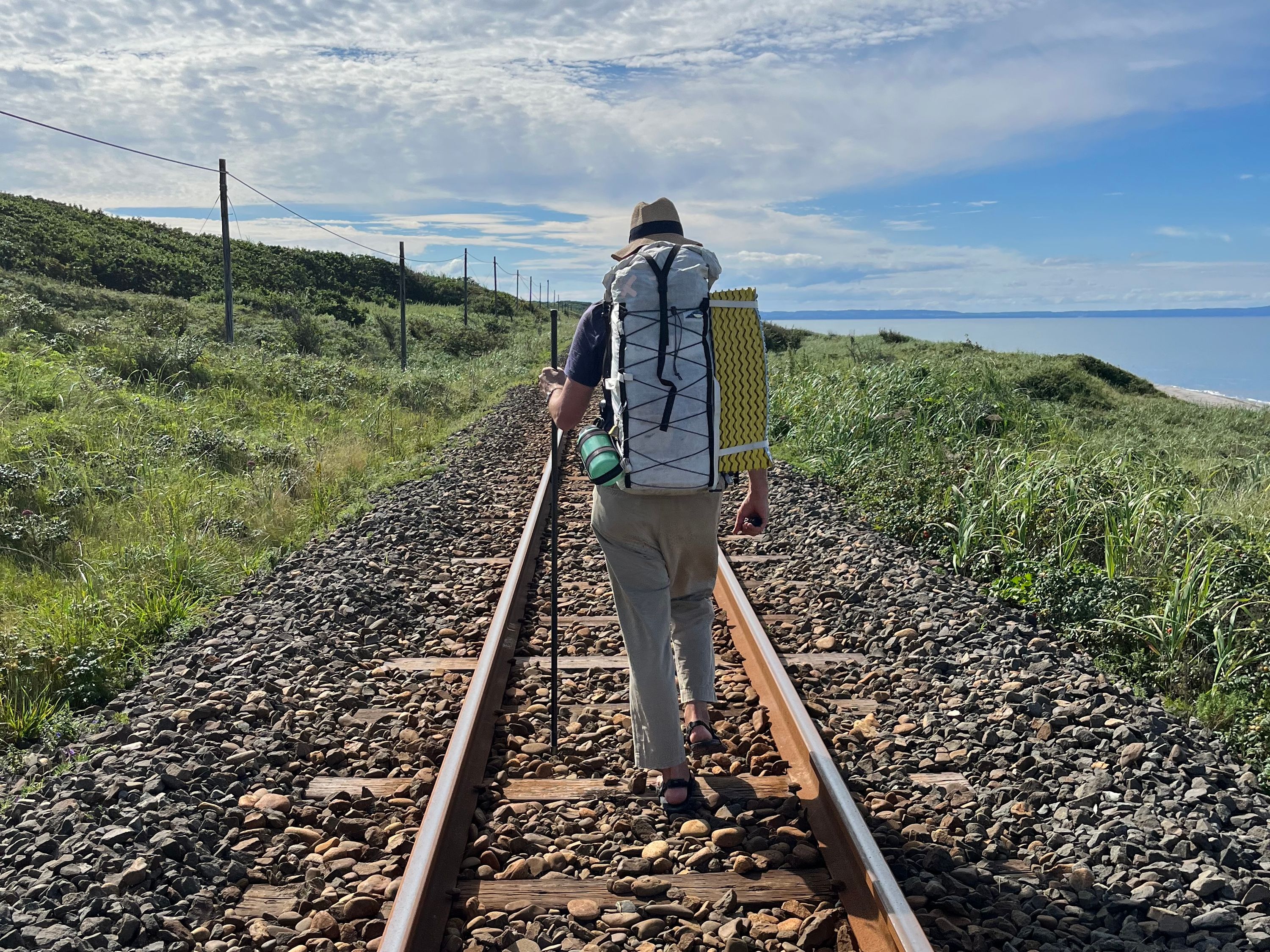 Peter Orosz in a large backpack walks away from the camera on a railroad track running by the sea.