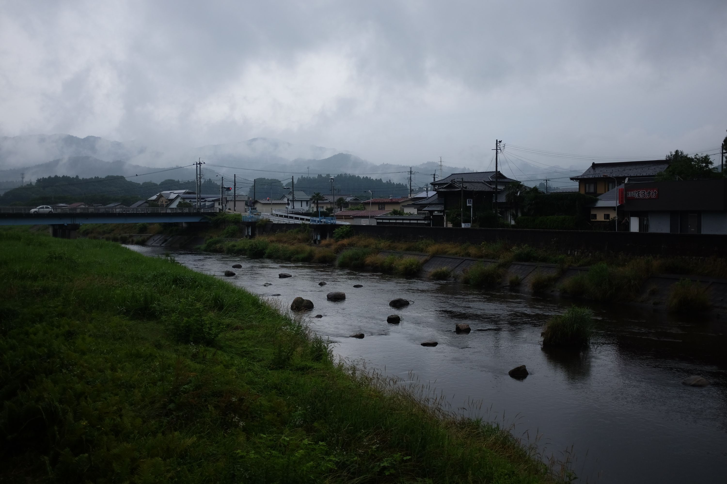 A village by a small river under overhanging grey clouds.