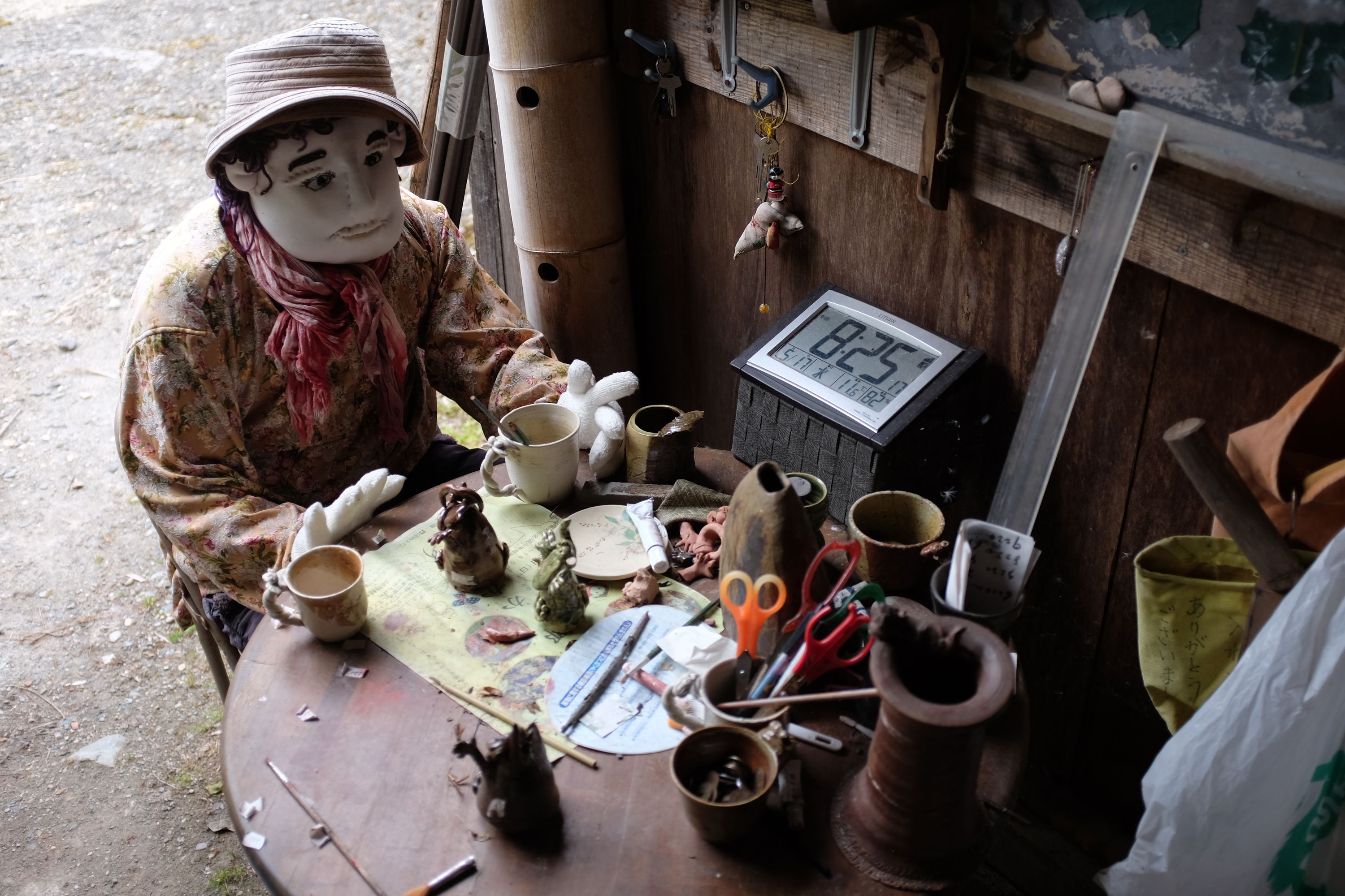 A doll sits at a low worktable with mugs and various tools laid out on it.