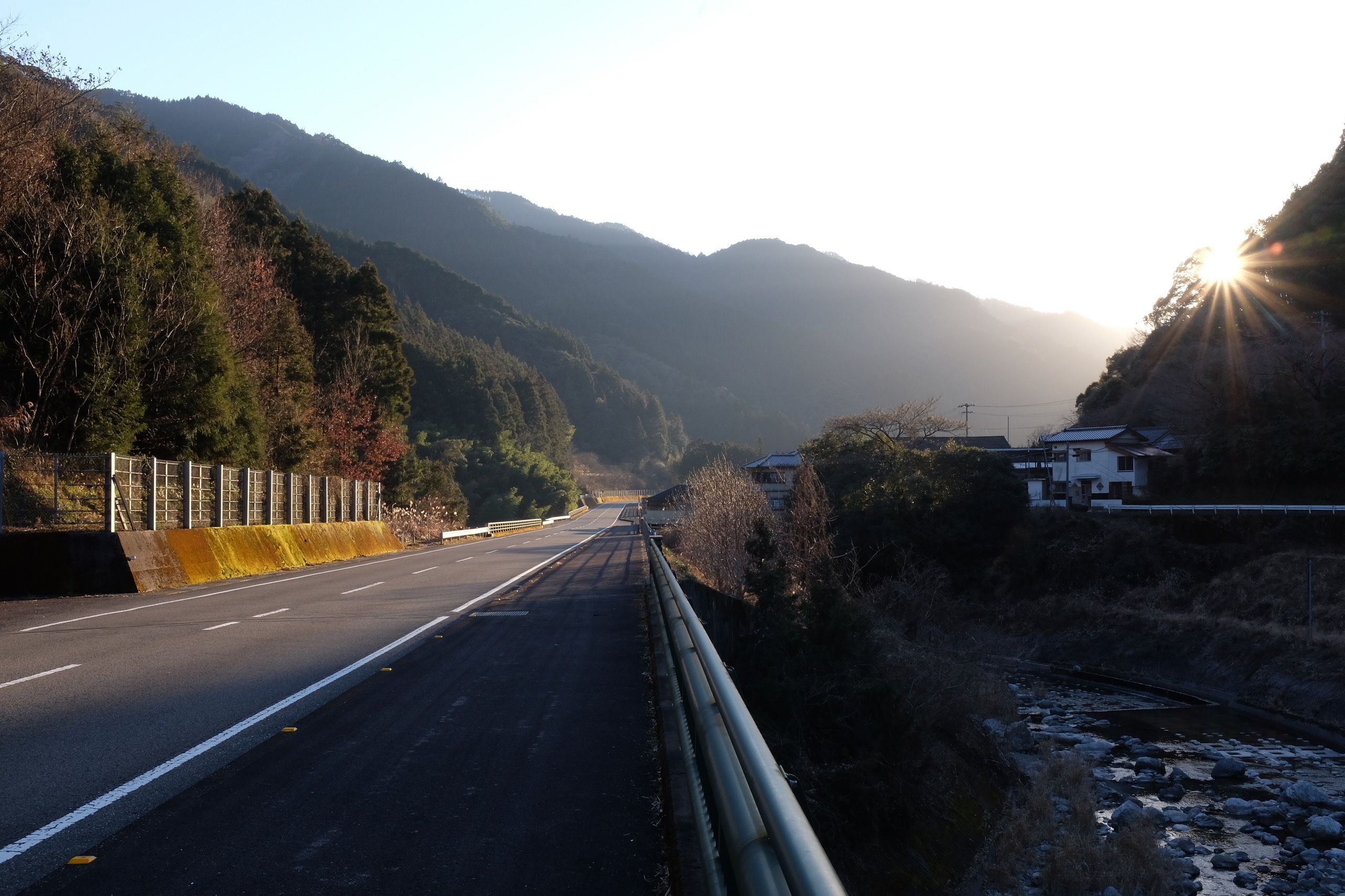 The sun sets behind the hills in a valley lined by low forested hills.