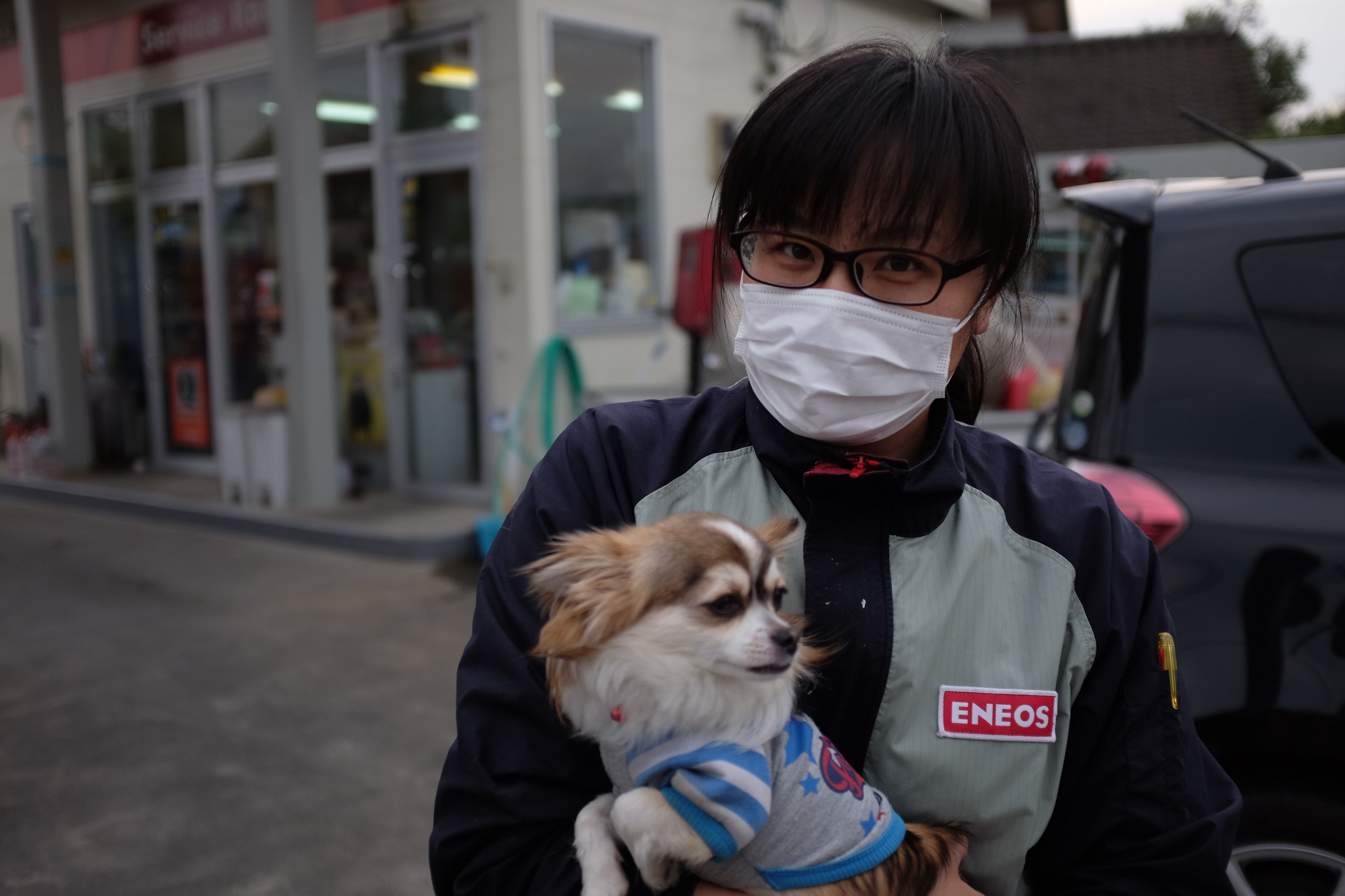 A woman wearing a surgical mask and mechanic’s overalls holds a small dog who wears a striped jumper.