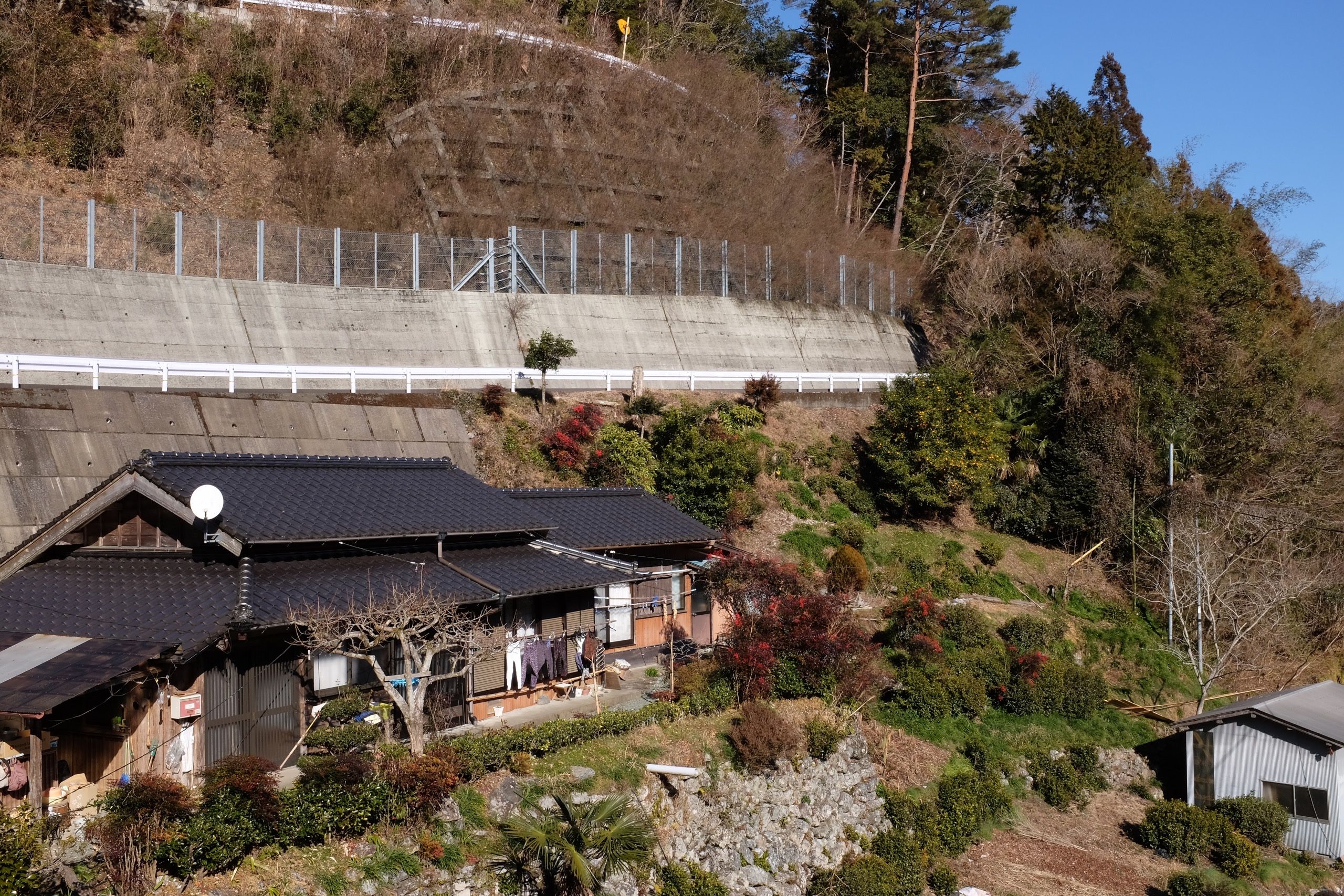 A small Japanese house on a hillside below a road.