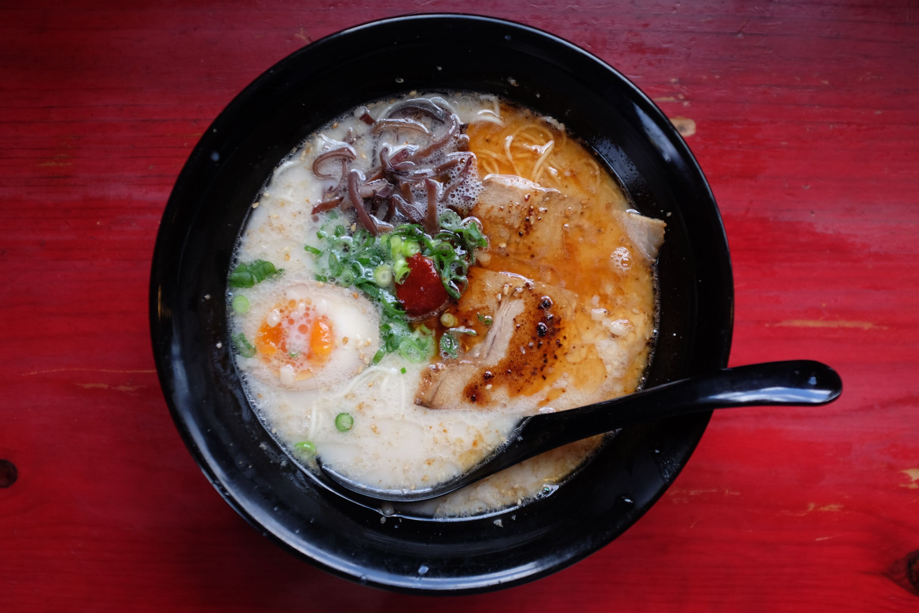 A bowl of white and yellow tonkotsu ramen on a red table, picking up the yellow of the Suzuki sign and the red of the car from the previous two pictures.