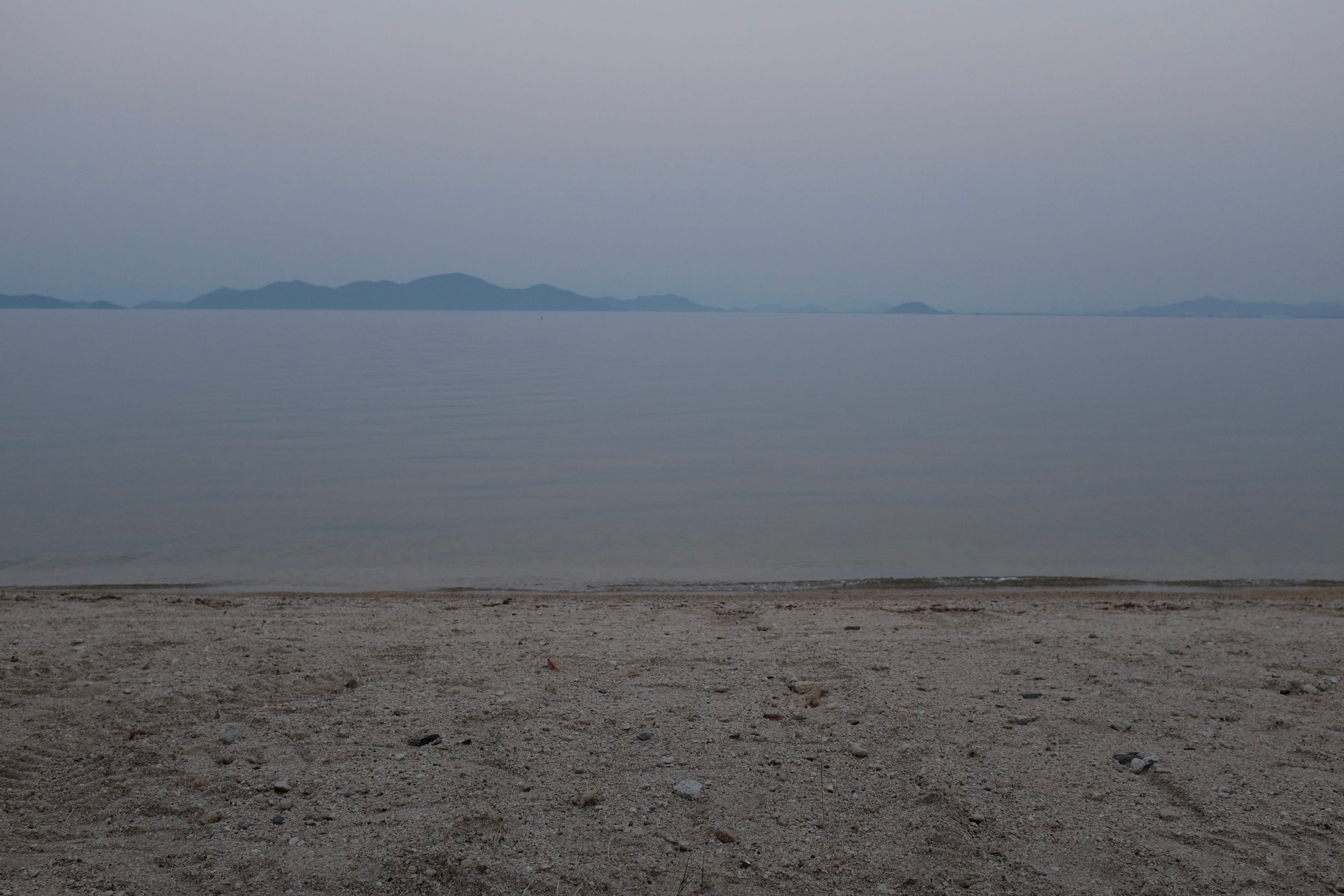 A sand beach almost the same color as the water and the sky, with hills on the opposite shore.