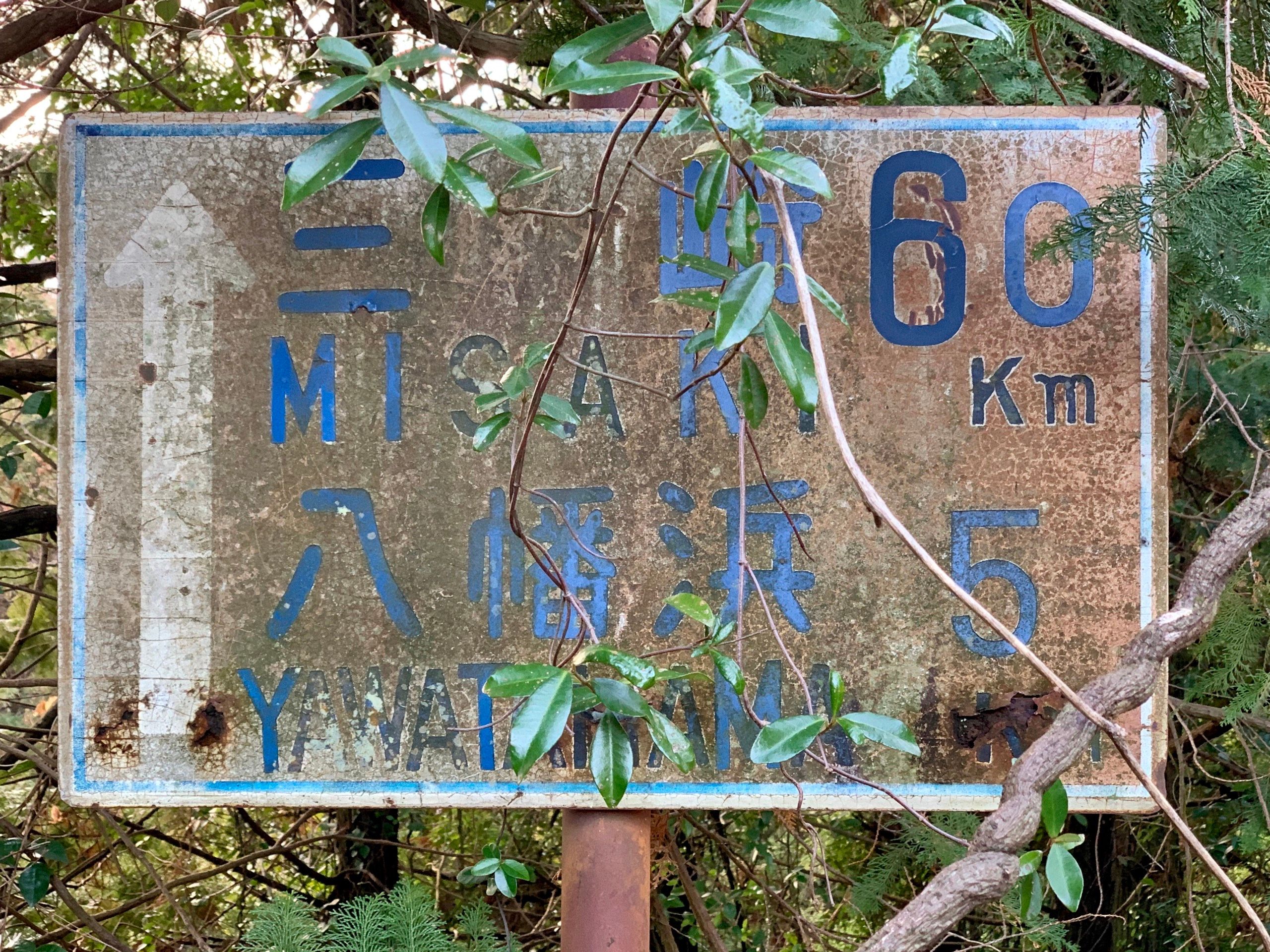 A rusty and overgrown sign shows the distances to Misaki (60 km) and Yawatahama (5 km).