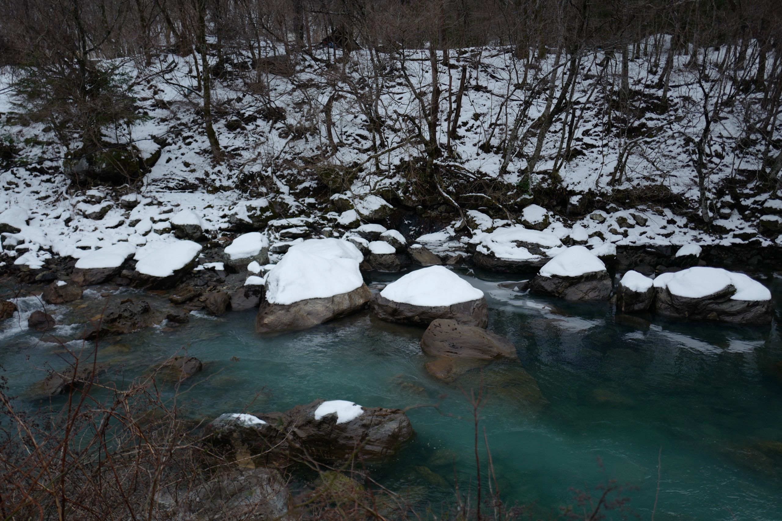 Snow-covered boulders in a turquoise-colored stream, with snow on its banks.