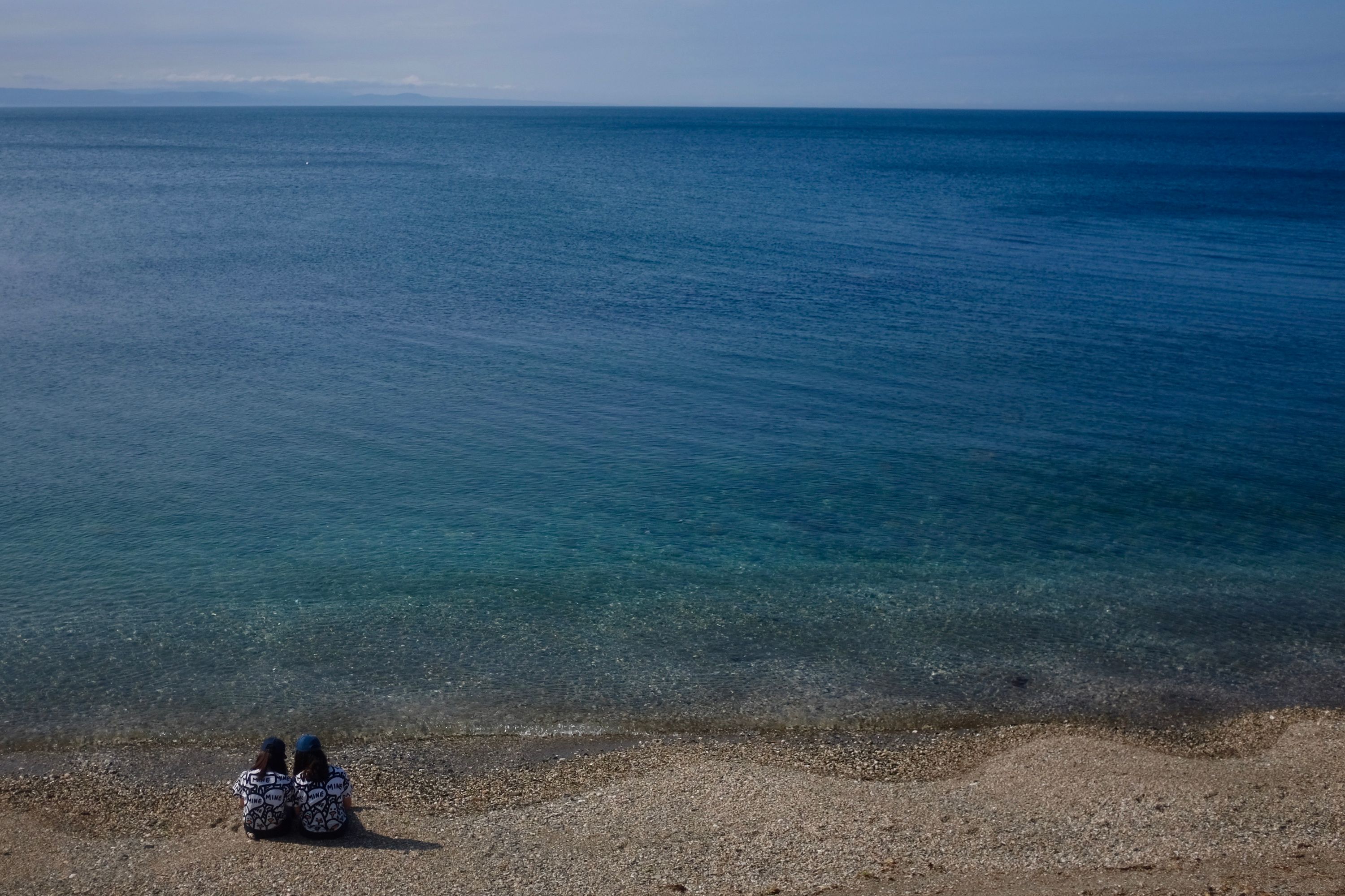 Two girls in identical clothes sit on the beach and look out on the sea, which looks vast compared to how small they are.