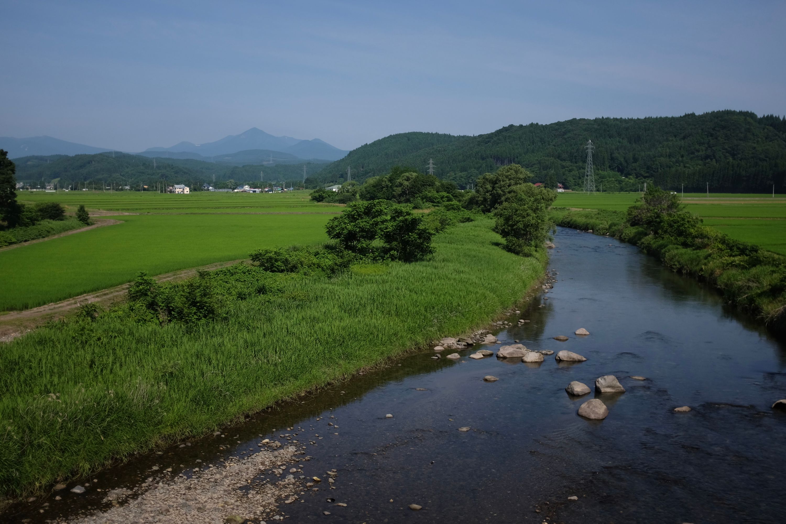 Beyond a river and green rice fields, the Hakkōda Mountains rise in the distance.