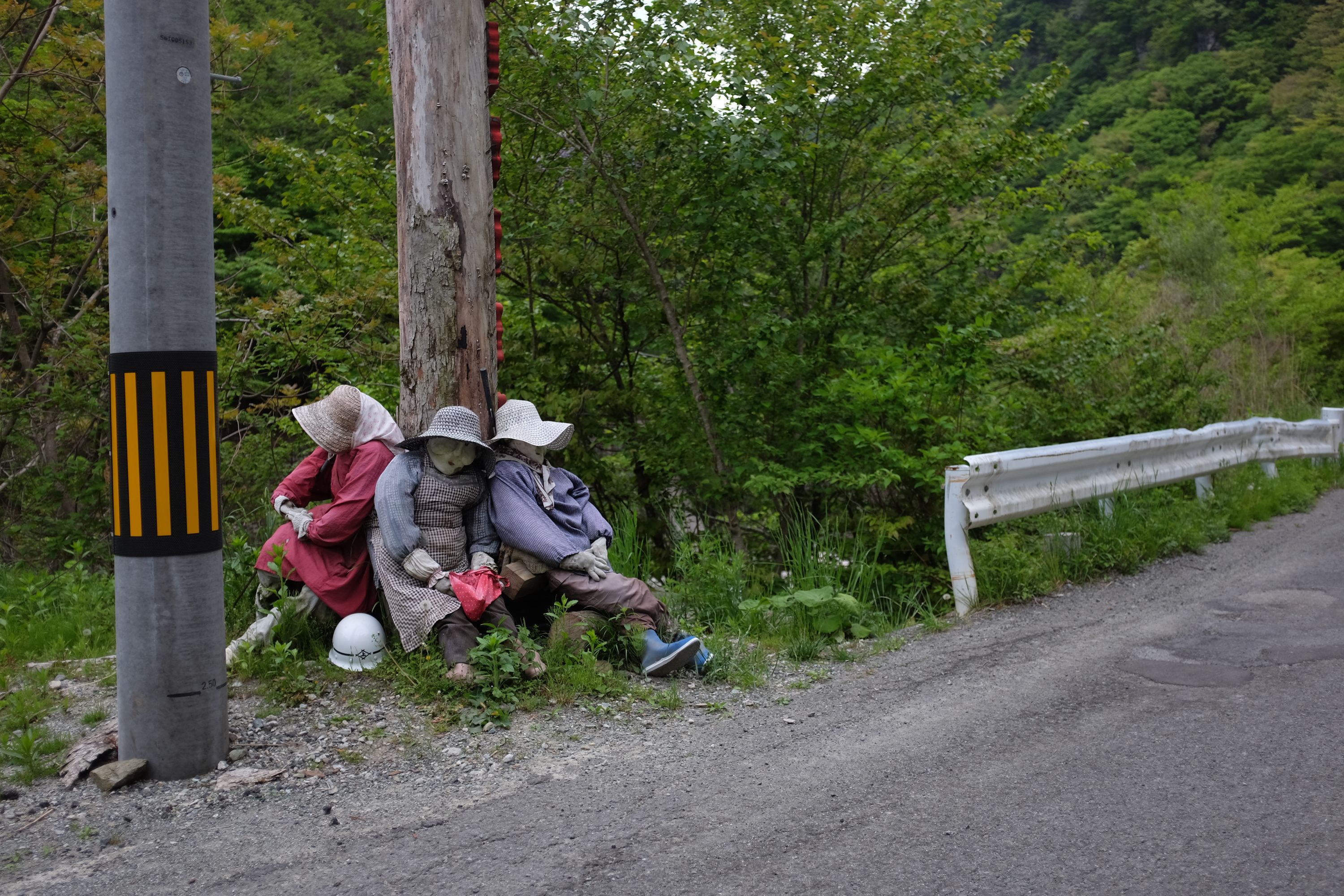 Three human-sized dolls dressed up as village women sit on the ground around a wooden telephone pole.