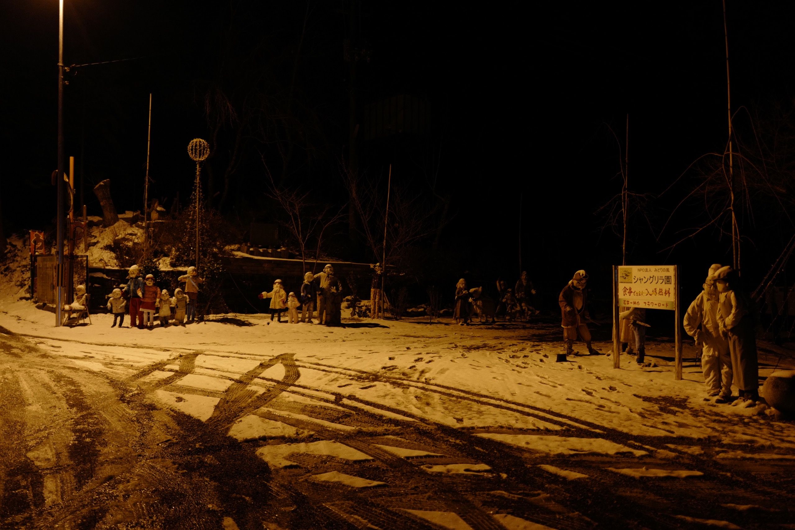About 20 human-sized dolls lounging at night in a snowed-in area by a narrow road.