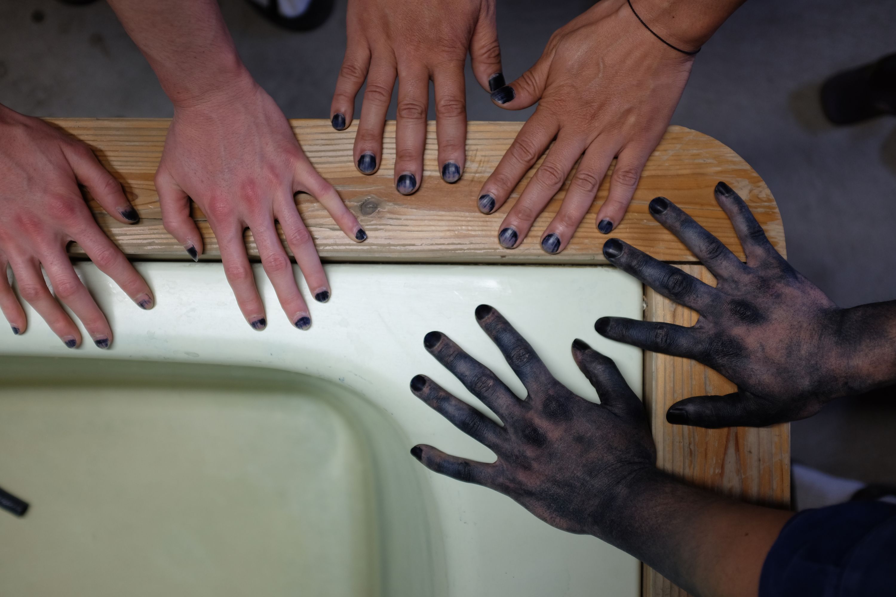 Three pair of hands, the fingernails of all dyed indigo blue, placed on the edge of an indigo dyeing vat.