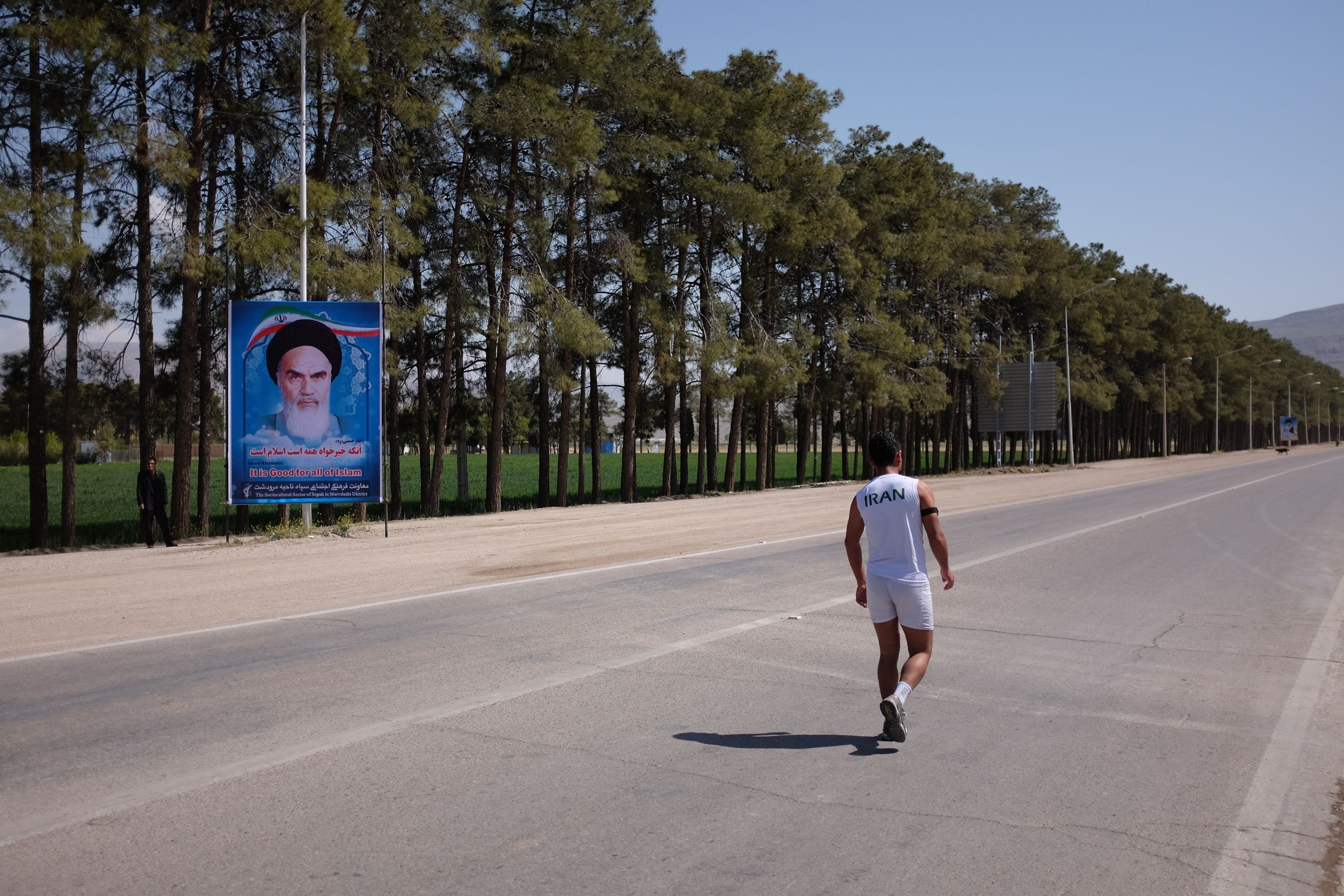 A man in white running clothes jogs past a picture of Ayatollah Khomeini on a very straight country road.