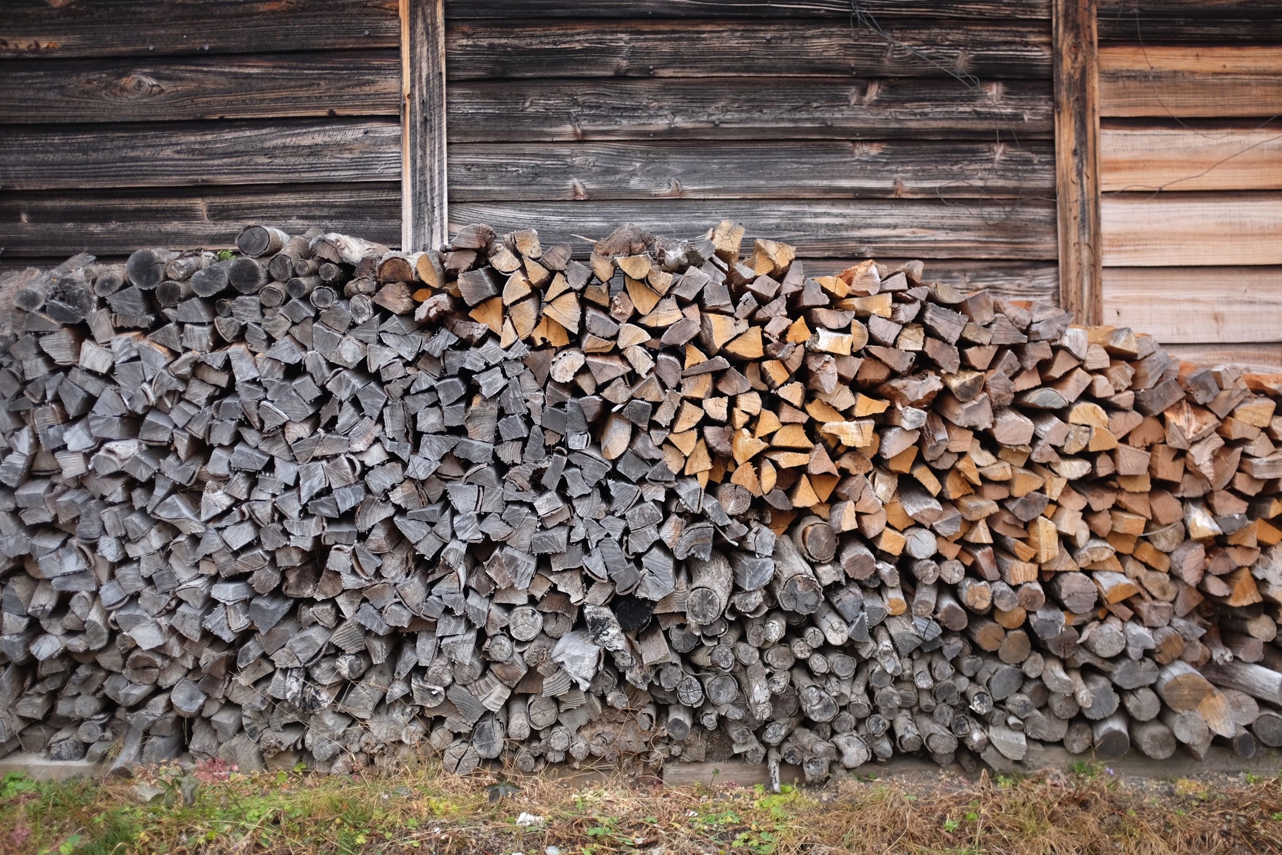 A heap of old and new firewood stacked against the wooden wall of a house.