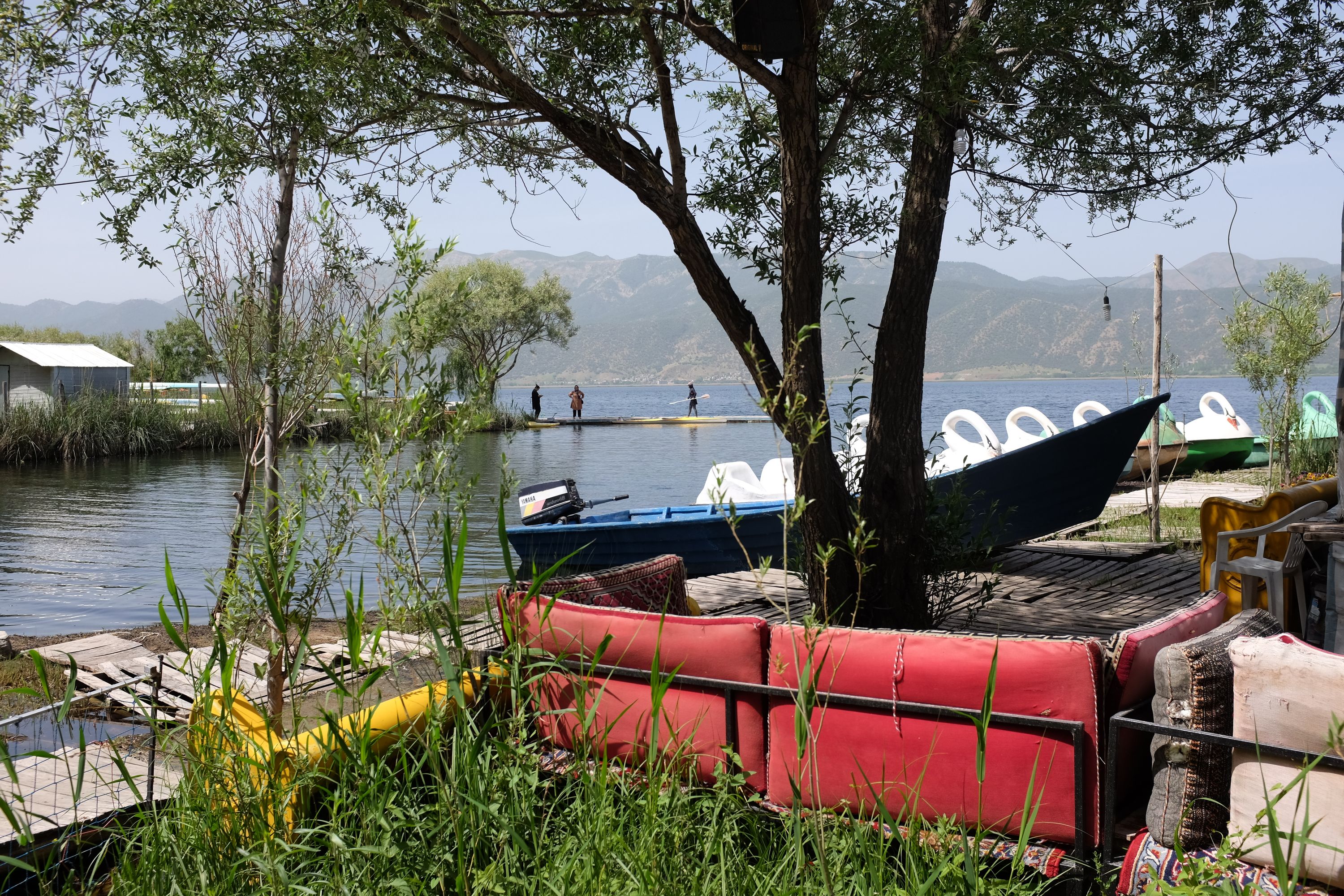 On a pier on a normal-looking lake, three women gear up to kayak, with white swan boats anchored near them.