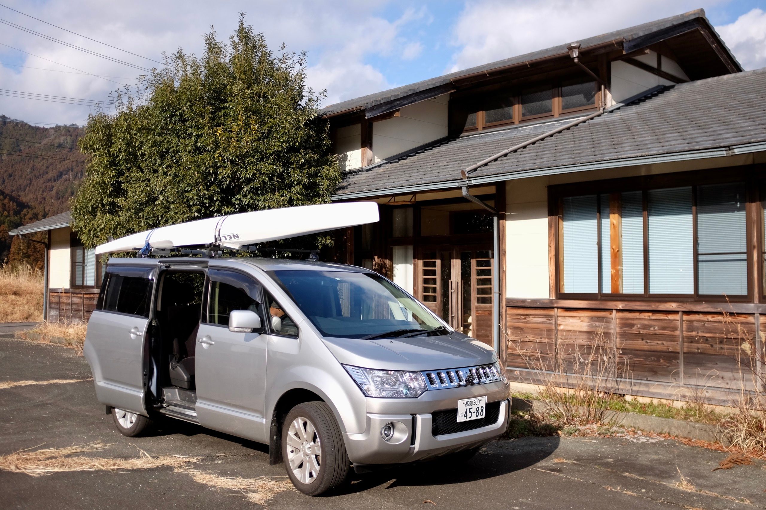 A minivan with a white racing kayak lashed to its roof parked in front of a wooden Japanese house.