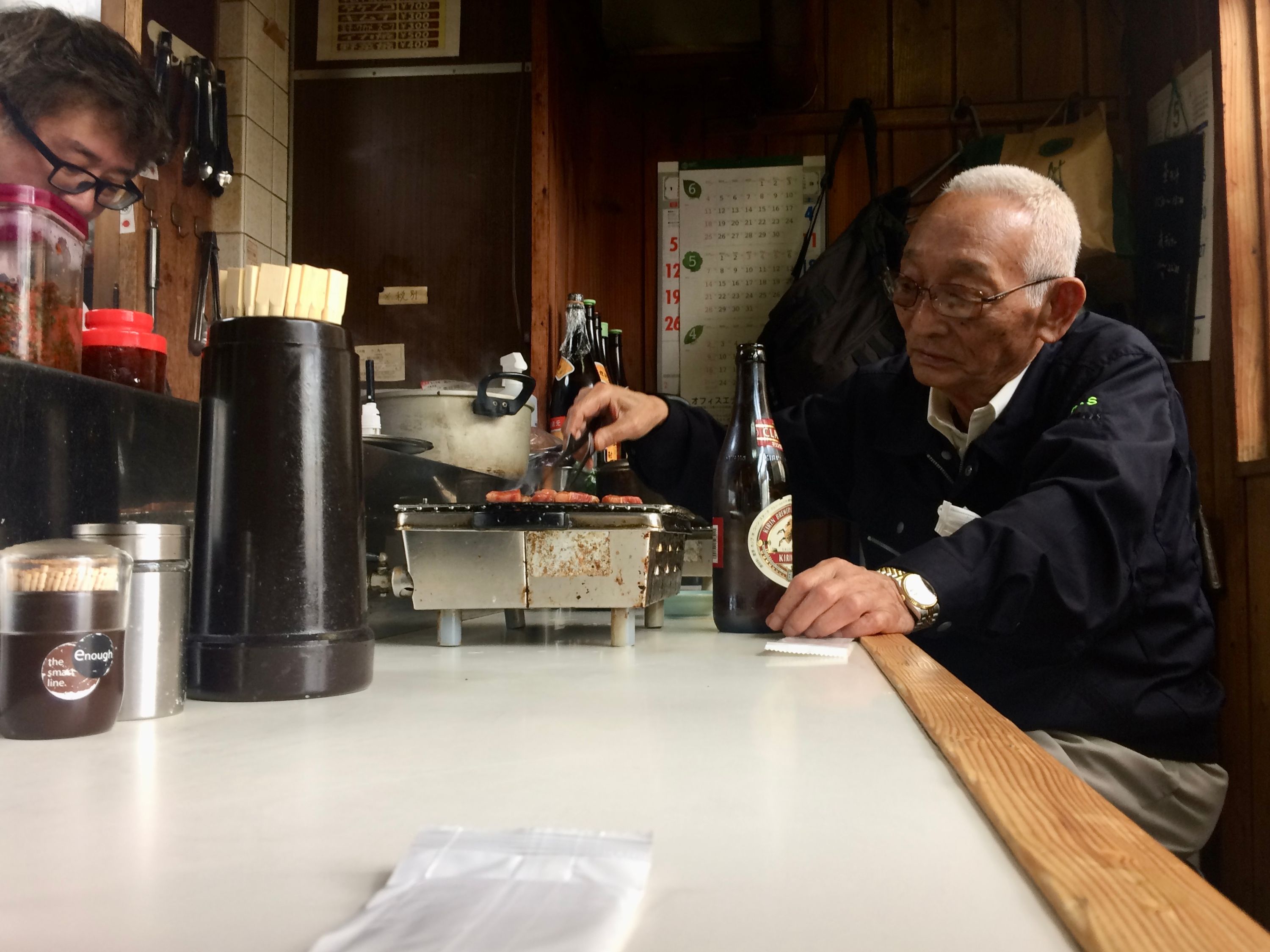 An elderly Japanese man grills pieces of Kōbe beef on the counter of a small restaurant, a bottle of beer near his left hand.