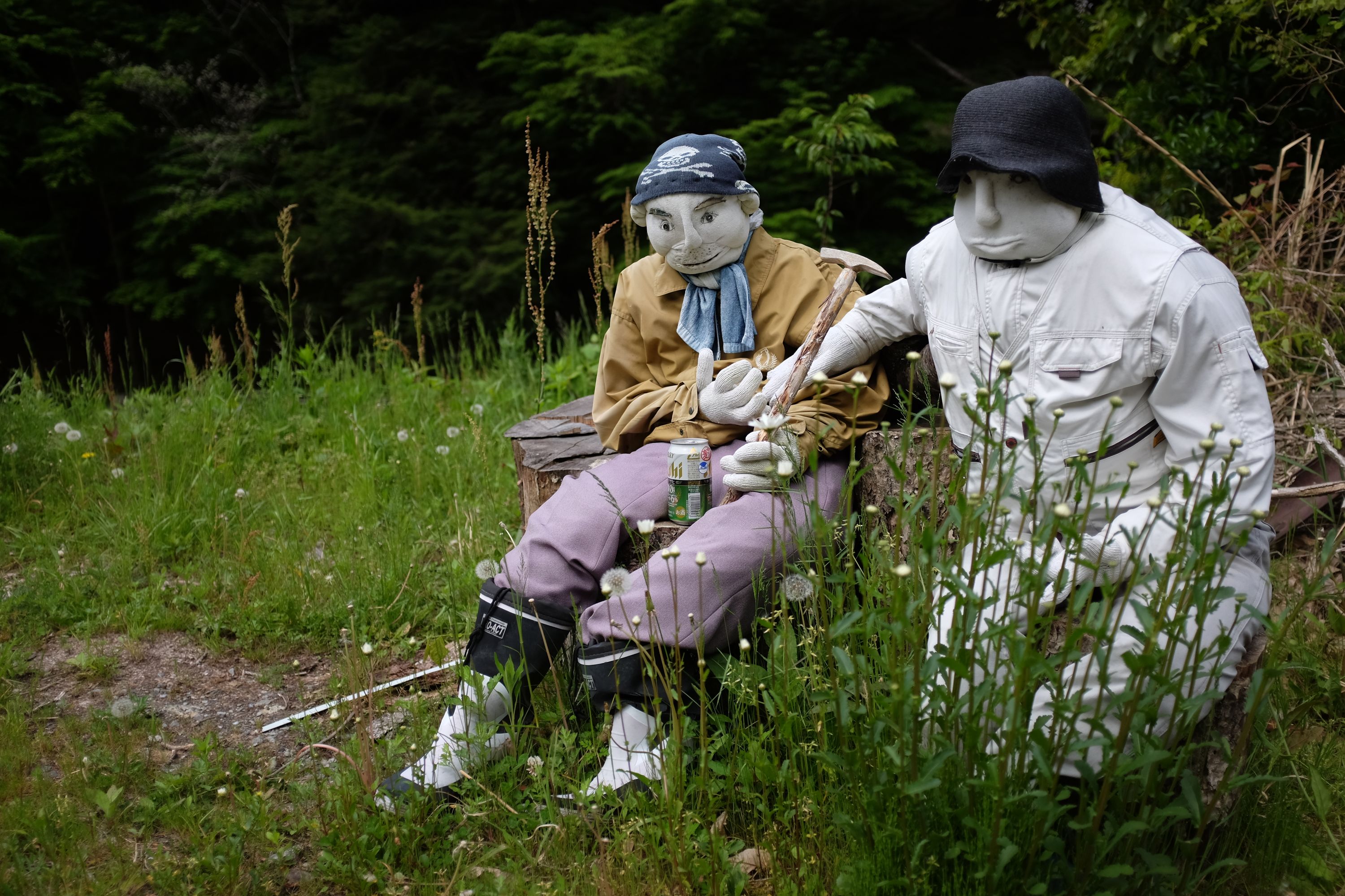 Two dolls sit on stools in a field, drinking beers.