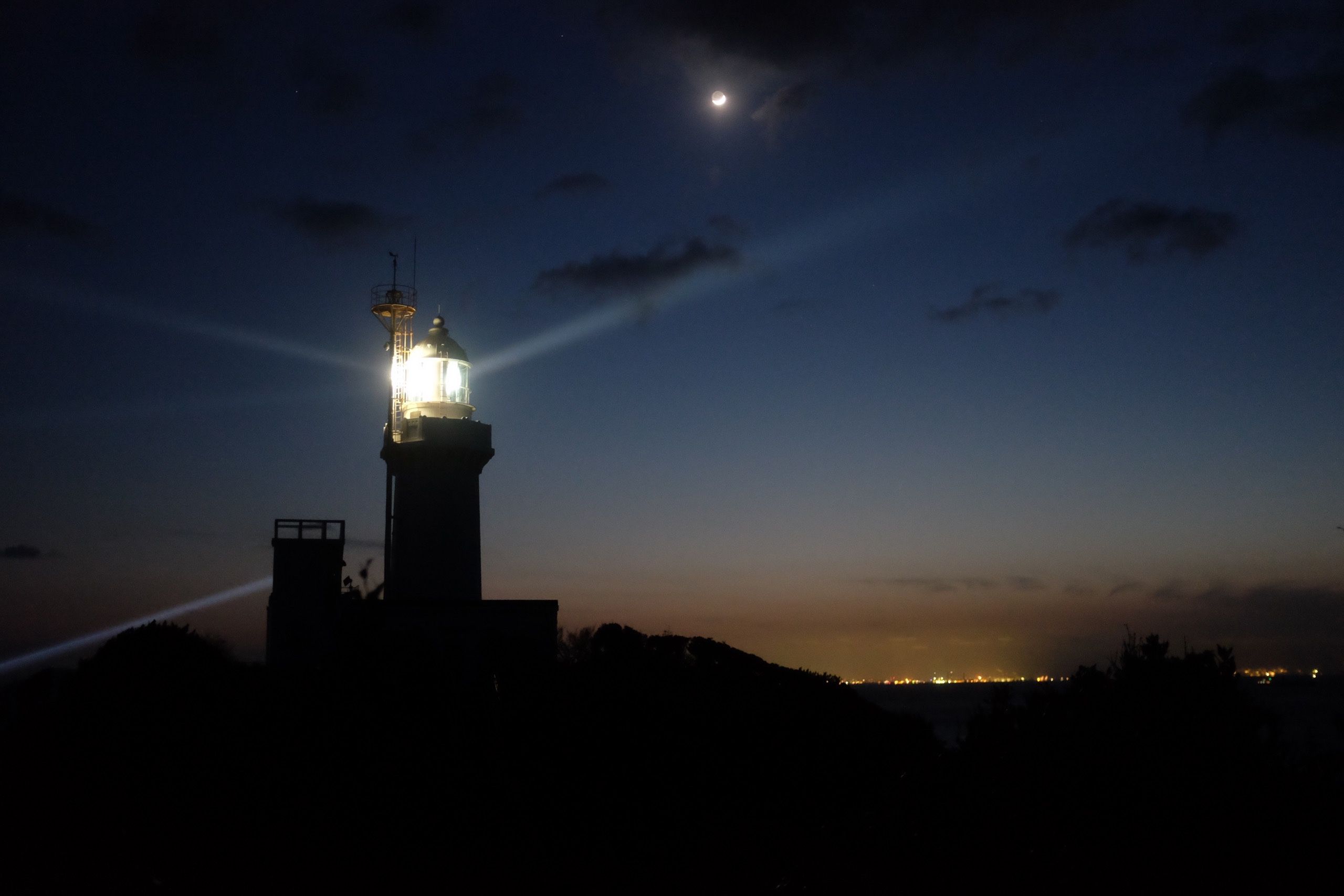 A lighthouse casts rays into the night sky. The lights of a city are visible on the horizon behind it.