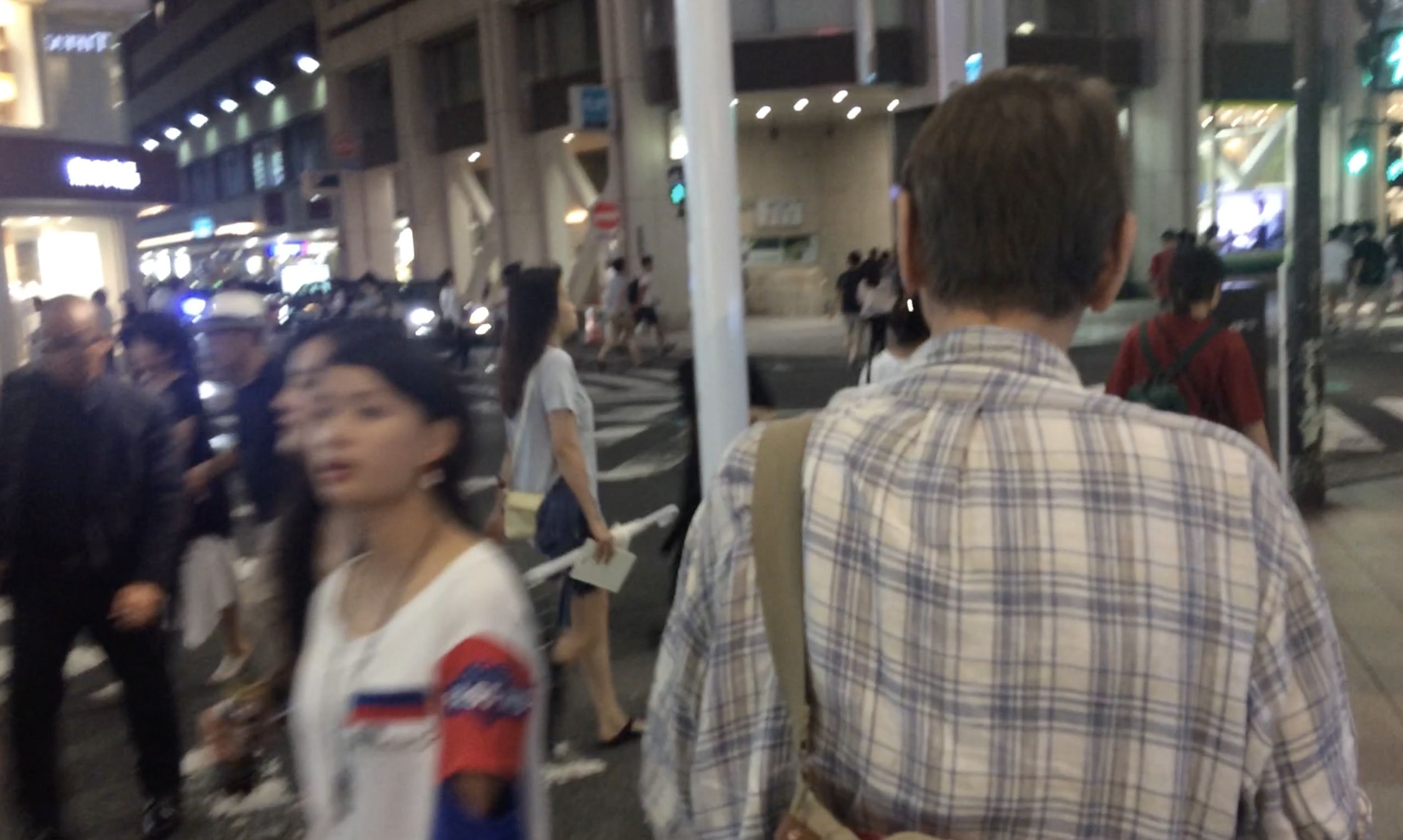A somewhat blurry nighttime photograph showing people in a busy urban area. In the foreground, the back of a man wearing a light-colored checkered shirt is visible as he walks away from the camera. The man is Timothy Harris. To his left, a young Asian woman in a white t-shirt with red and blue graphics is visible in profile, also walking. The background shows the bustling atmosphere of a Japanese city at night with bright lights, store signs, crowds of people, and what appears to be a busy pedestrian area or station entrance. The motion blur and lighting create the dynamic feeling of Tokyo’s nightlife.