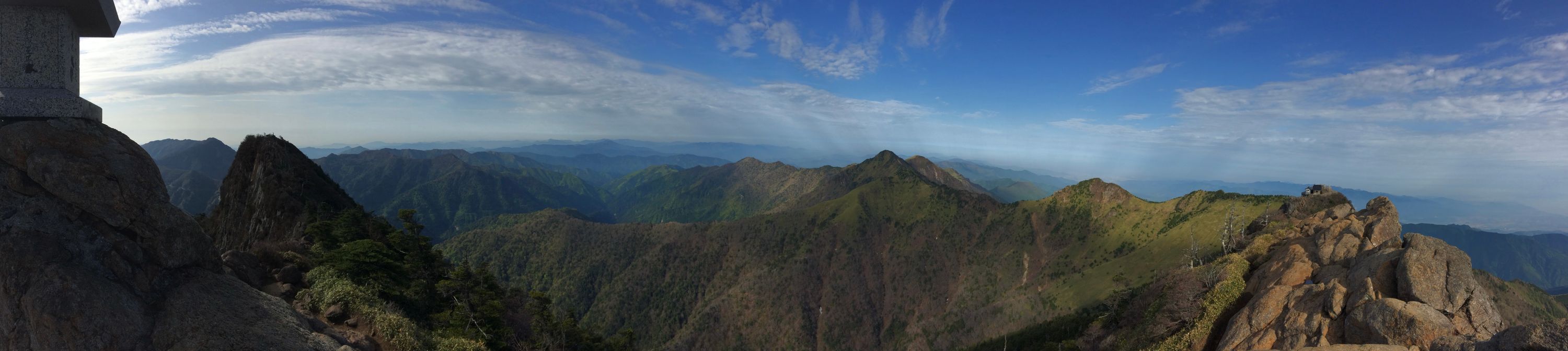 A very wide panorama of jagged mountains under a blue sky streaked with cirrus clouds, taken from the summit of Mount Ishizuchi looking north-northwest.