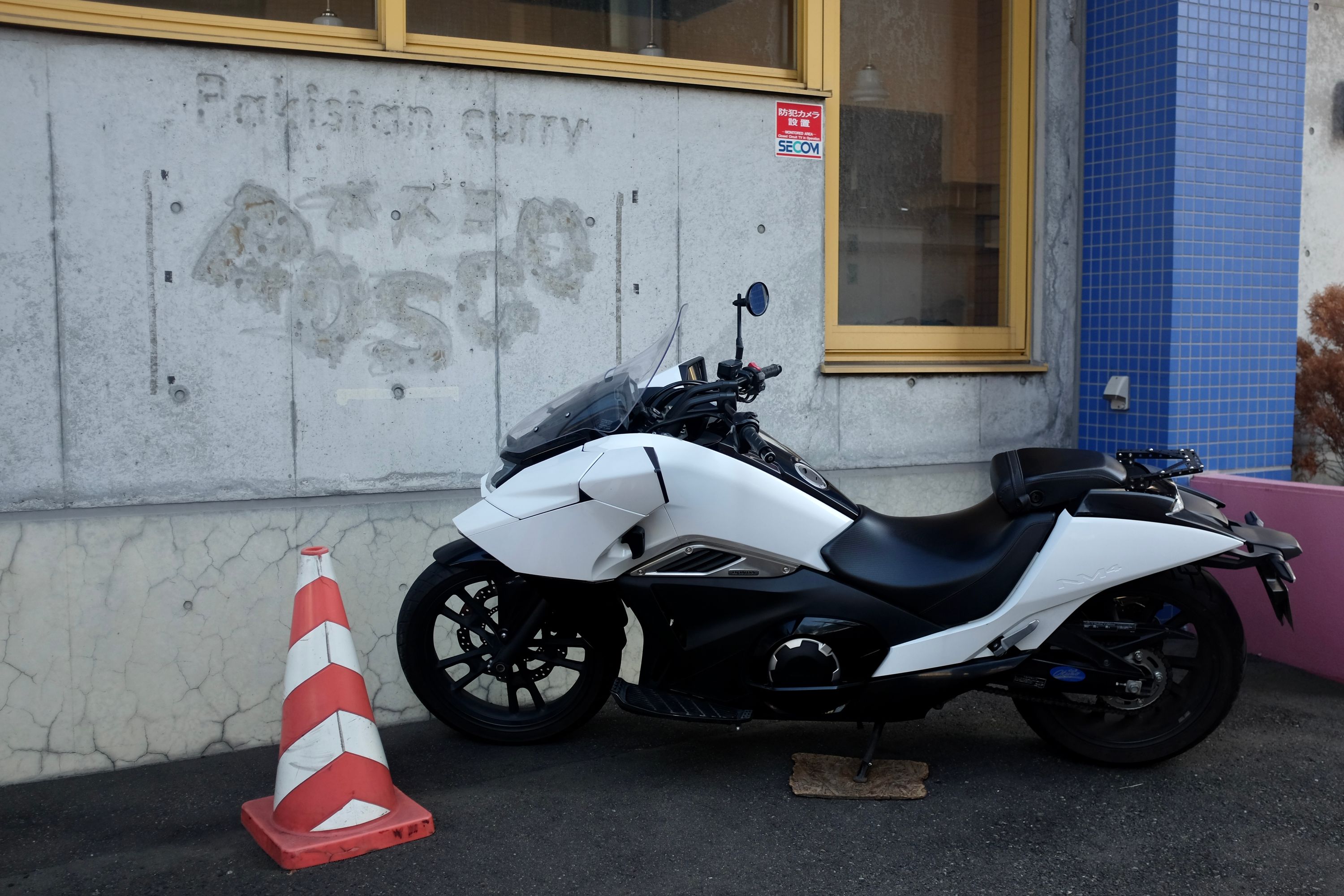 A futuristic-looking white motorcycle parked in front of a concrete wall with the faded sign “Pakistan curry”.