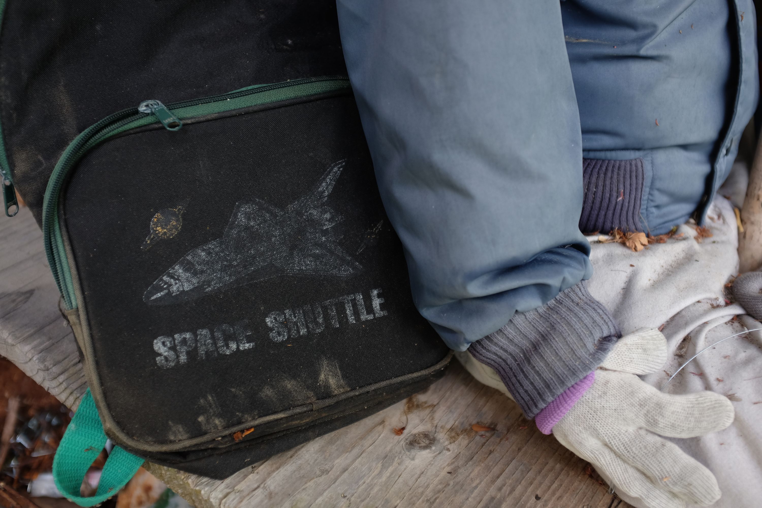 An old canvas briefcase with a picture of the Space Shuttle is placed next to the right hand of a doll.