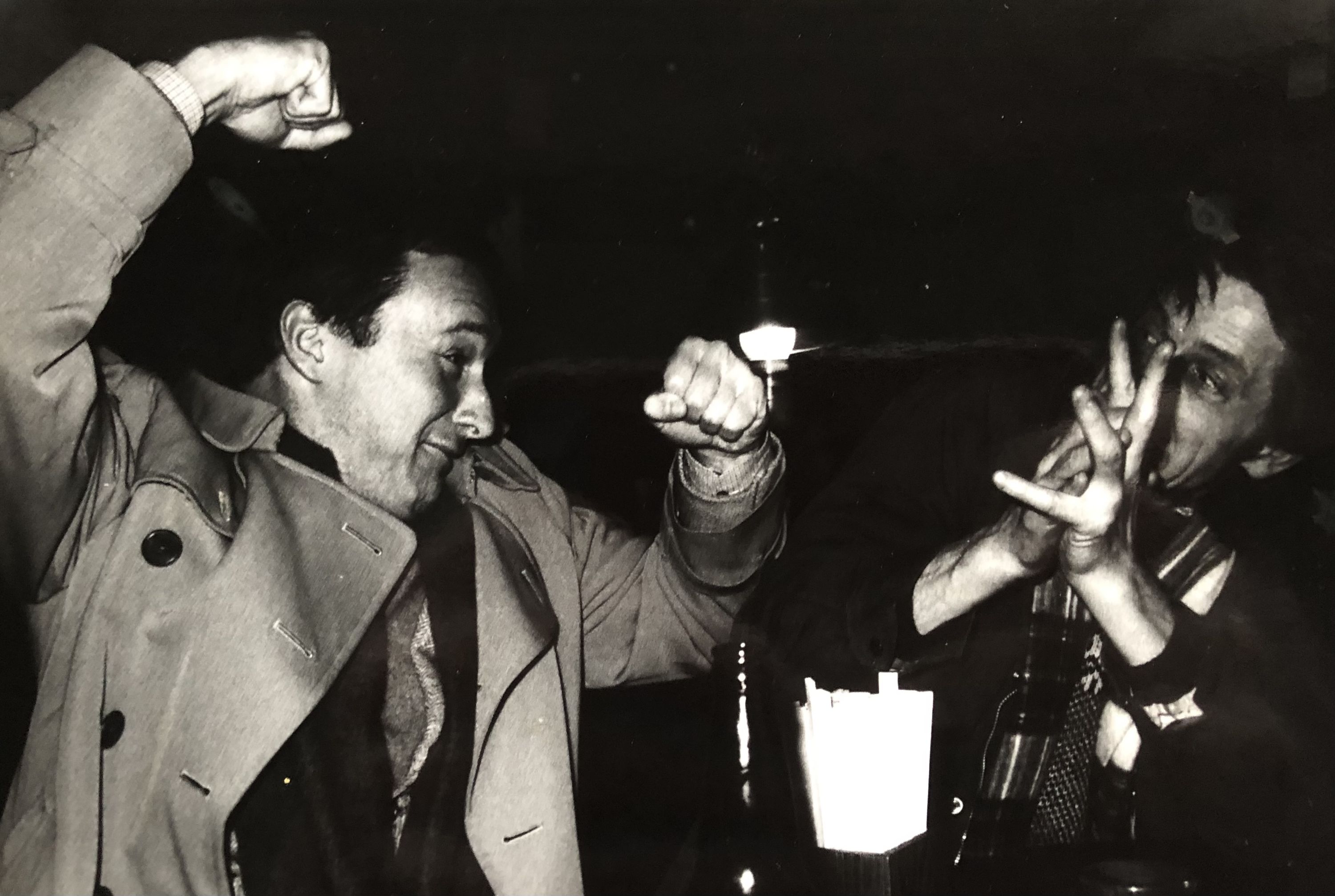 A black and white photograph of two men celebrating in a Tokyo bar in the 1980s. On the left, Alan Booth wears a light-colored trench coat and raises his right fist triumphantly in the air. On the right, Timothy Harris grins widely with both hands raised near his face. The scene captures a moment of joyful camaraderie, with the atmospheric lighting and composition typical of casual bar photography from that era.