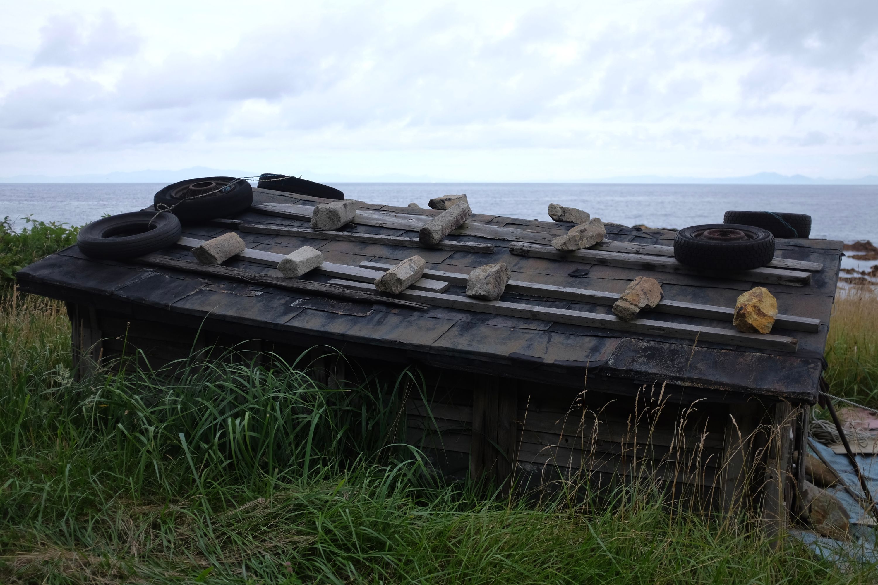 A wooden shack on the shore has its roof weighed down by rocks and car wheels.