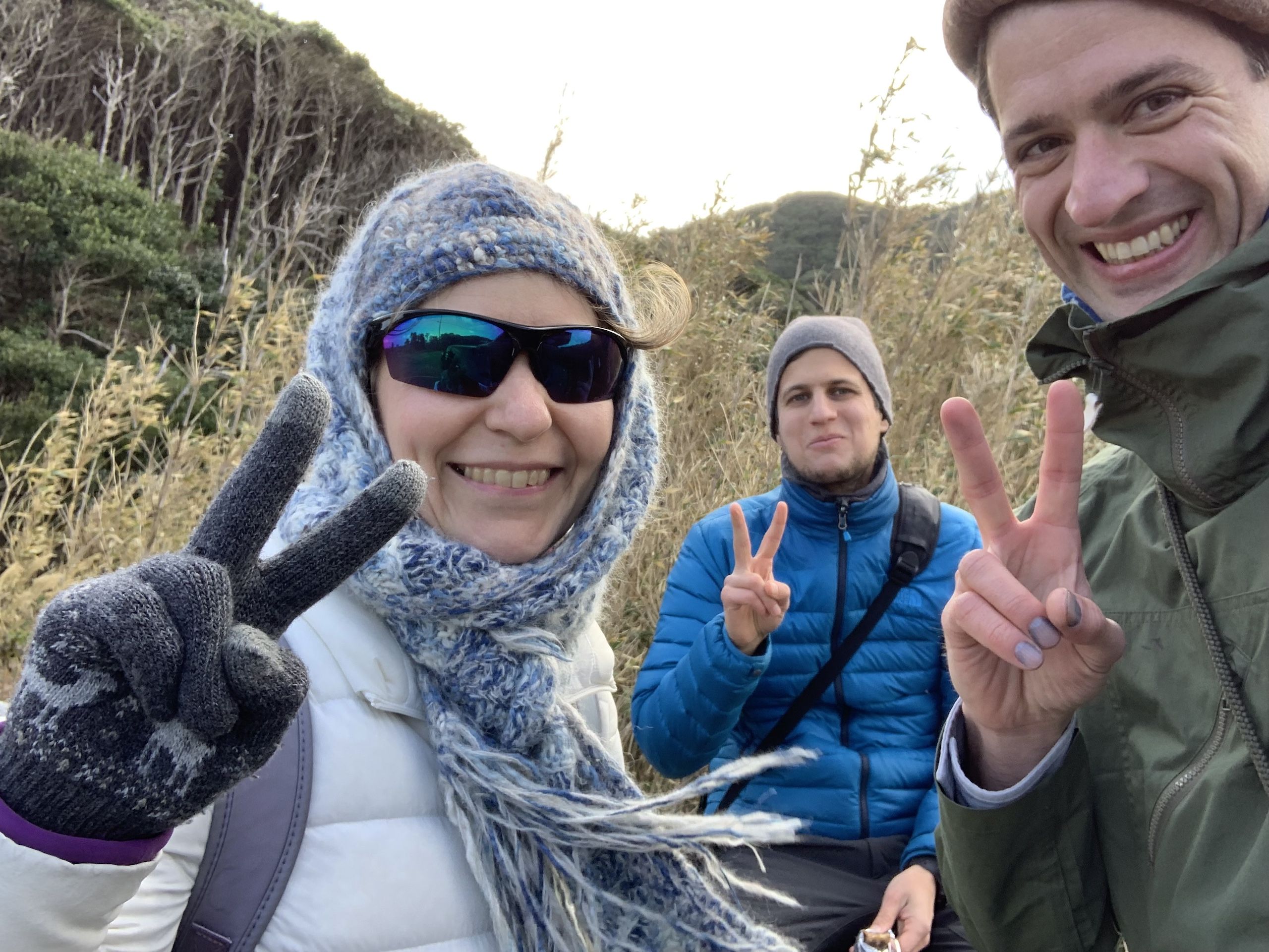 A color photograph taken outdoors showing three people in hiking gear making peace signs with their hands. On the left is Hanga Yoshihara-Horváth, a woman wearing sunglasses, a blue and white patterned head scarf, dark gloves, and a white jacket with a purple backpack strap visible. In the center is Gyula Simonyi, a man wearing a gray knit beanie and a bright blue puffy jacket, also making a peace sign. On the right is Peter Orosz, a man wearing a brown flat cap and an olive green jacket, holding up two fingers in a peace sign while smiling broadly. Behind them is wild grassland and hills covered with bare trees, suggesting late fall or early spring weather. The lighting appears to be natural daylight, creating a cheerful atmosphere of friends enjoying an outdoor adventure.