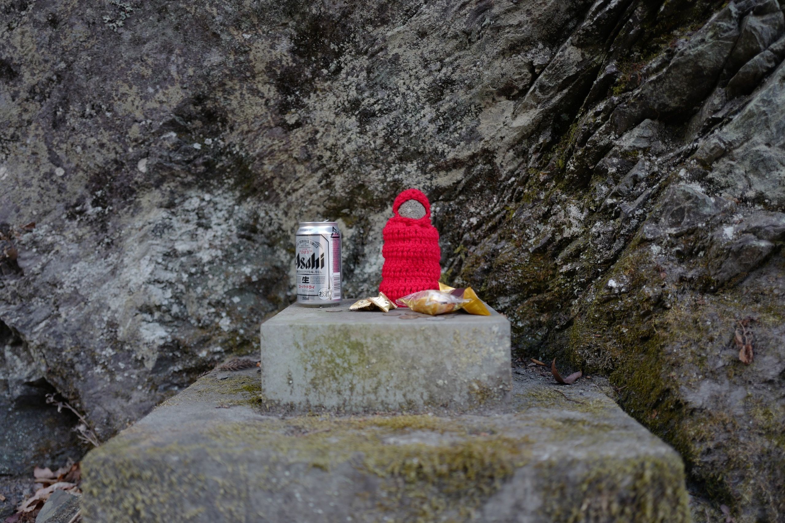 A small roadside deity in a red jumper, about the size of a can of beer, stands on a shrine next to an actual can of beer.