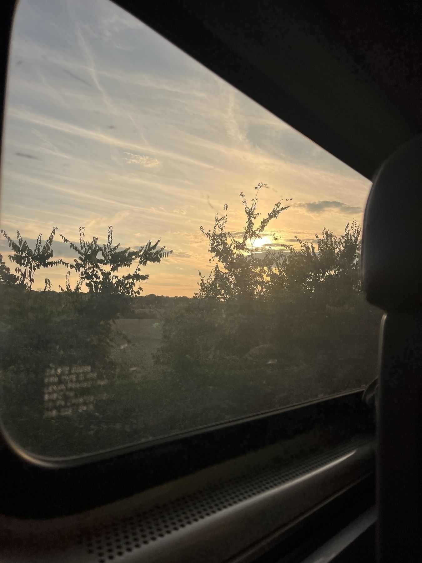 Looking out of a train window at a sunset with light blue skies, streaks of white cloud and a warm peach glow