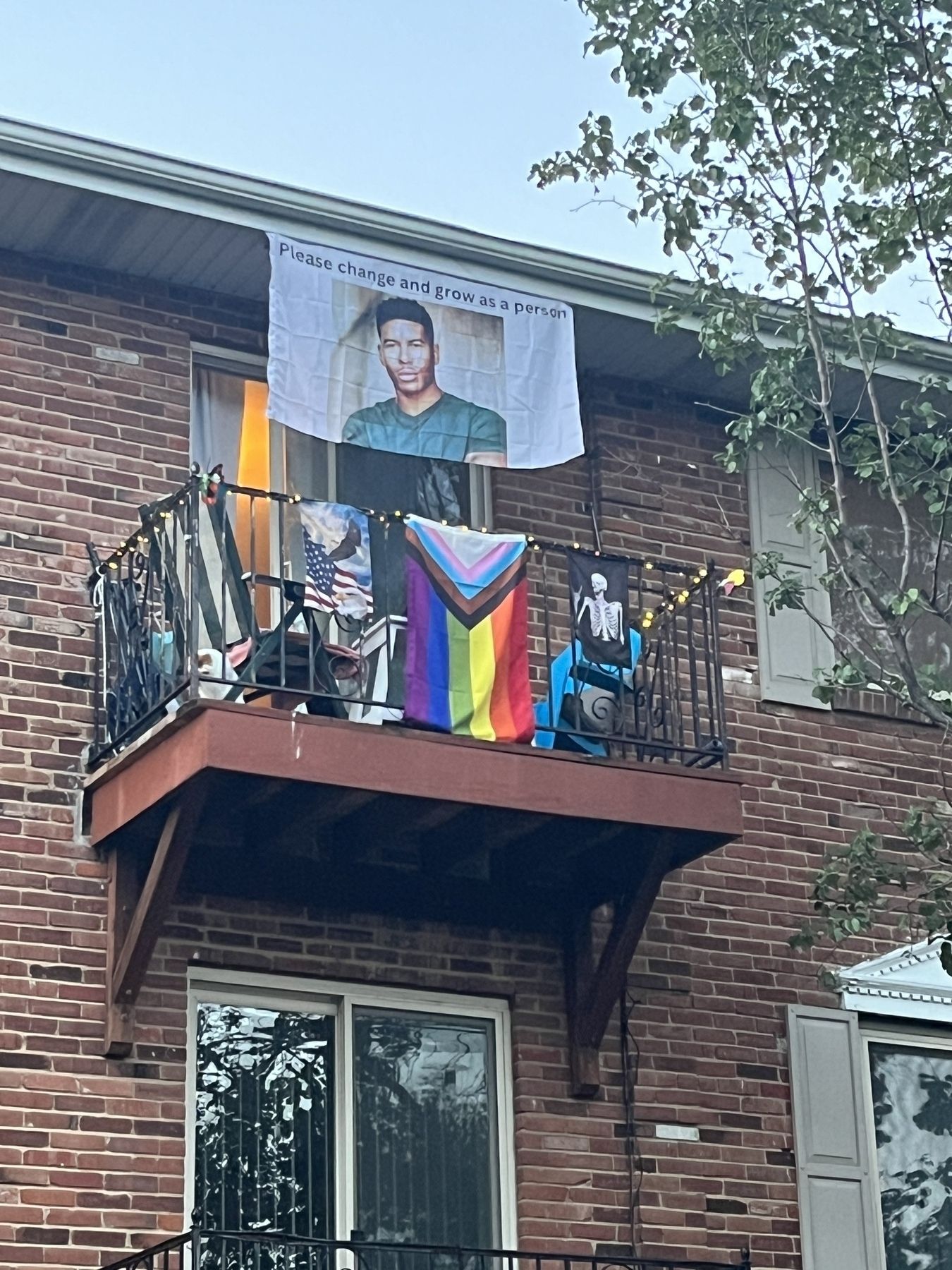 A brick apartment building with a Balcony with four flags - pride, skeleton rocker, America eagle, and a man saying please change and grow as a person