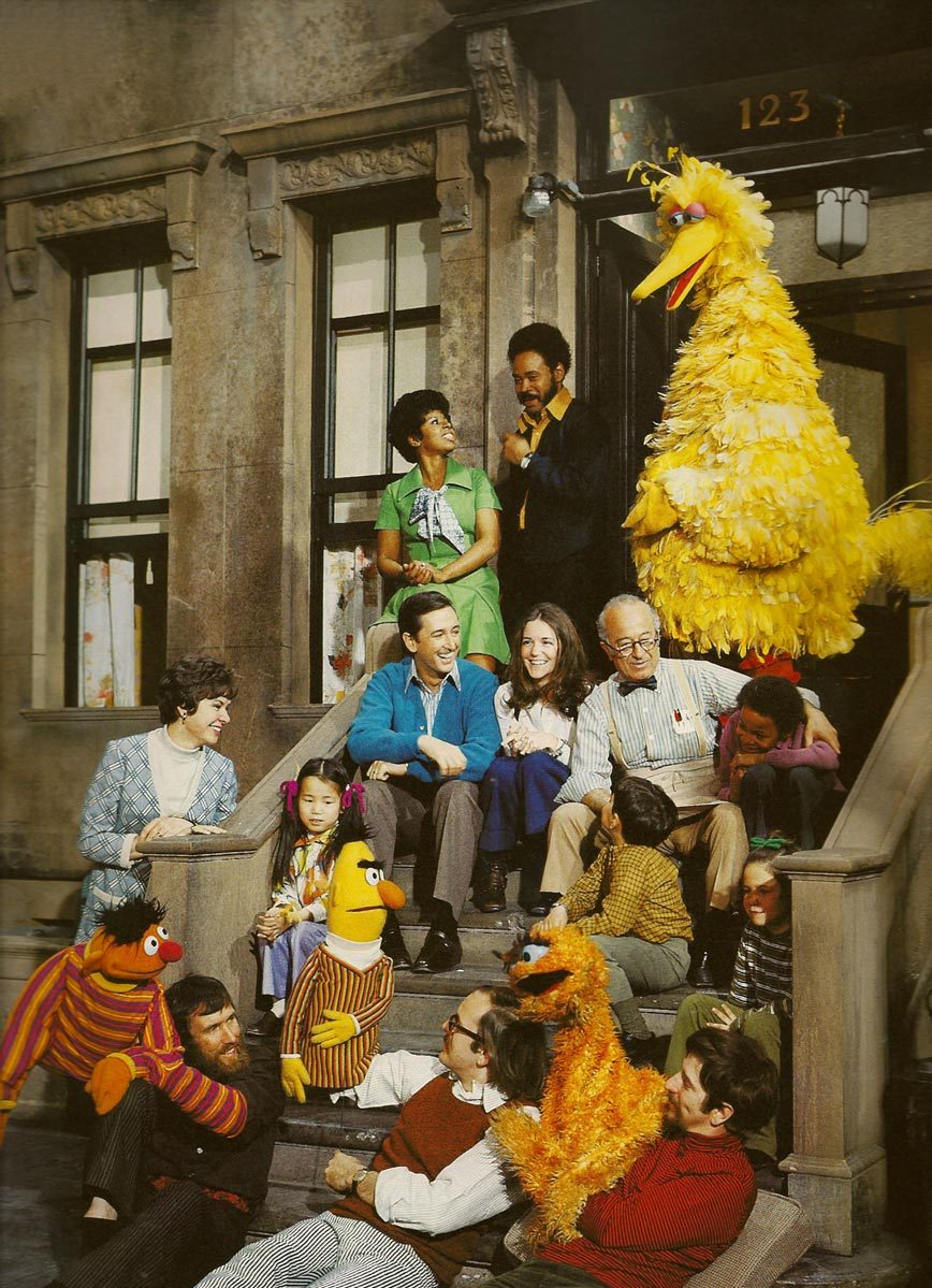 Characters of Sesame Street from the early years, including the Muppets, all sitting on the stairs and stoop of one of the sets